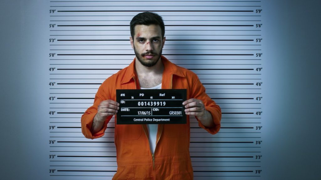 In a Police Station Arrested Man Getting Front-View Mug Shot. He's Wearing Prisoner Orange Jumpsuit and Holds Placard. Height Chart in the Background. Shot with Dark Cold Lights, Vignette Filter.